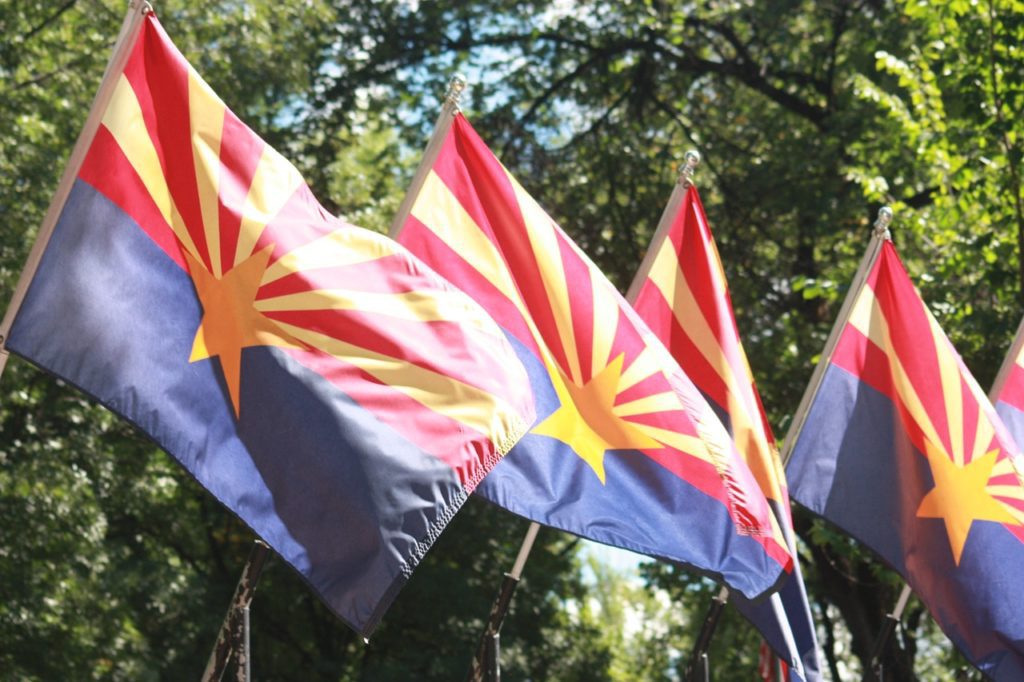 A row of Arizona state flags with a backdrop of trees on a bright, windy day.