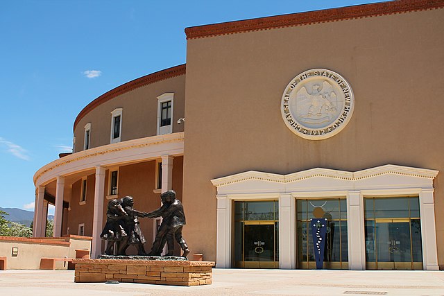 A photograph of the New Mexico State Capitol in Santa Fe during the day. The Great Seal of the State of New Mexico can be seen above the entryway.