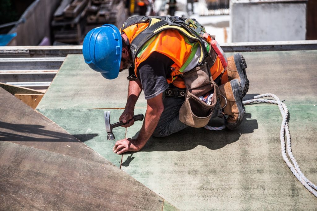 A construction worker wearing a hard hat and tool belt kneeling on a roof, hammering in a nail.