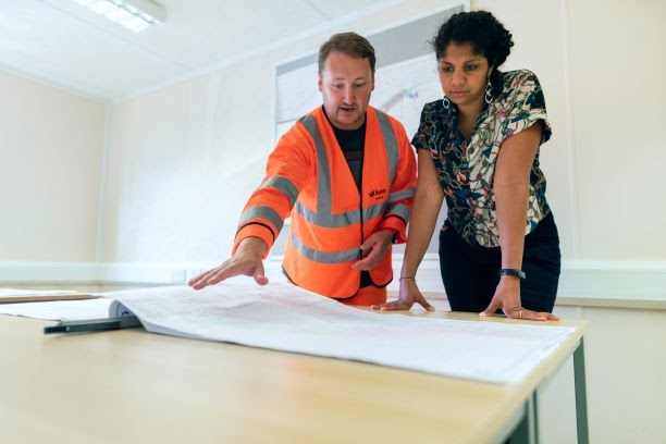 Two individuals, one in an orange reflective safety vest, the other in business casual attire, look over an architectural blueprint together in a white conference room.