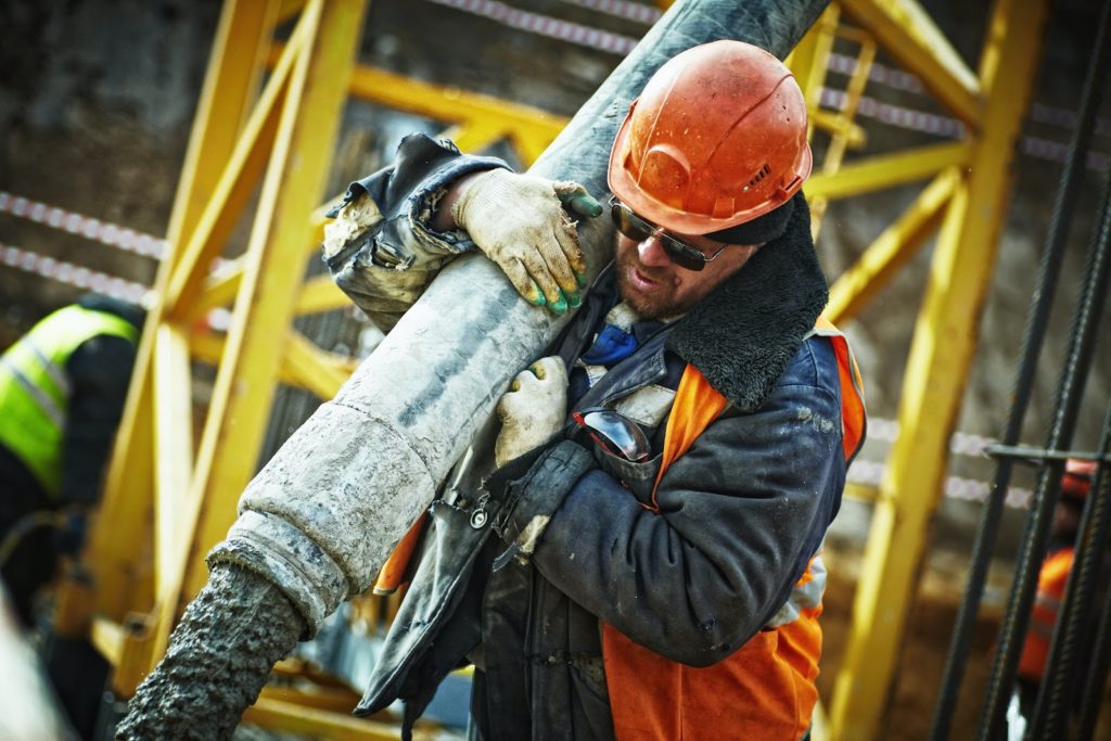 An individual in a hard hat, gloves, and orange safety vest pouring concrete.