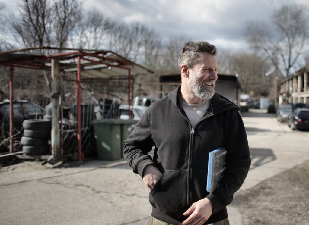 A contractor smiles while holding a clipboard, walking through a construction lot.