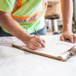 An individual in a reflective construction safety shirt makes notes on a clipboard atop a table, which is covered with architectural drawings.