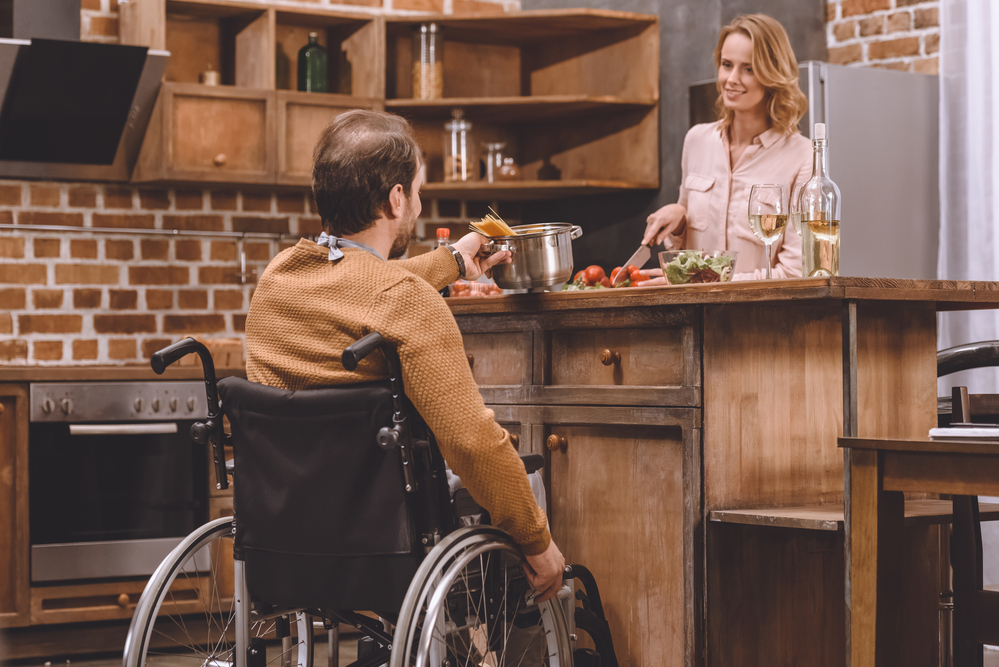 An individual in a wheelchair is seated next to an accessible center island in a domestic kitchen, placing a pot of spaghetti noodles on the counter. Standing on the opposite side, another individual uses a knife to chop vegetables.