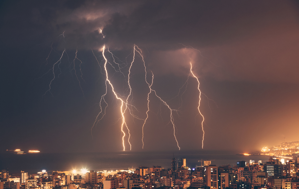 A lightning storm streaks through the cloudy evening sky in the background, with a large city skyline visible in the foreground.
