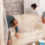 Couple wrapping sofa in plastic foil for protection while they are renovate living room. Home during renovation, decoration and painting. Interior apartment improvement maintenance. Roller, ladder for house repair