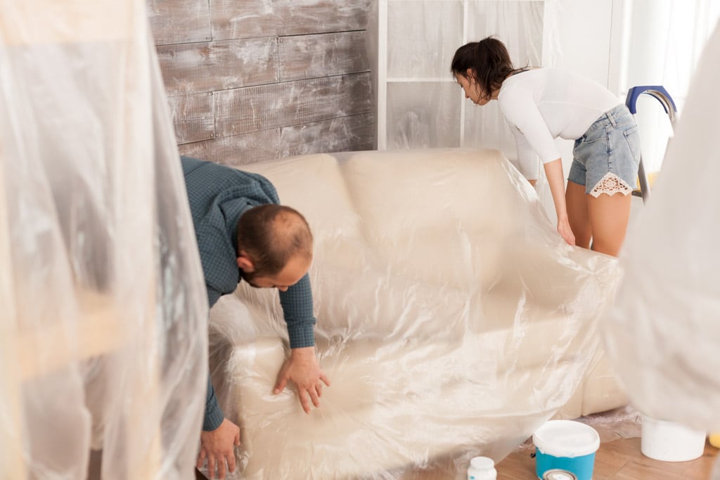 Couple wrapping sofa in plastic foil for protection while they are renovate living room. Home during renovation, decoration and painting. Interior apartment improvement maintenance. Roller, ladder for house repair